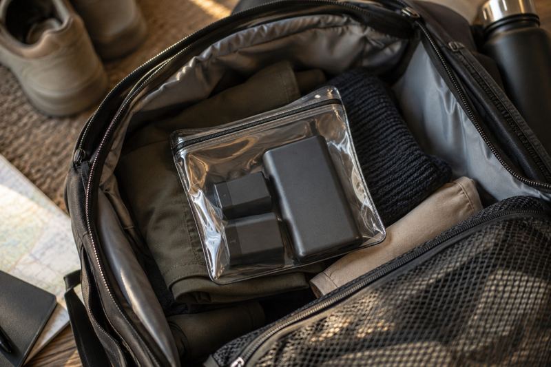 Spare camera batteries and a power bank in a clear pouch inside an open carry-on bag.