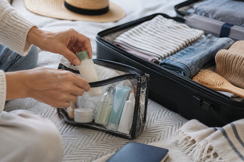 Hands packing travel-size bottles into a clear zip toiletry bag beside an open suitcase.